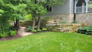 Stone wall and walkway leading to house with bright green grass and many trees.
